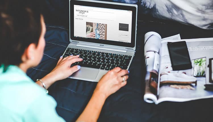 Woman browsing on a laptop next to magazines on a bed.