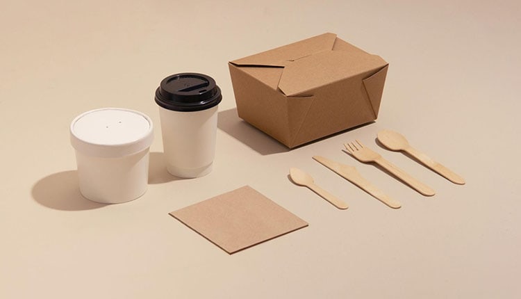 A takeaway packaging set on a light background, including a cardboard box, two paper cups with lids, wooden cutlery, and a napkin.