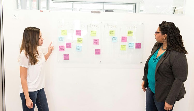 Two women discussing colorful sticky notes on a whiteboard in an office setting.