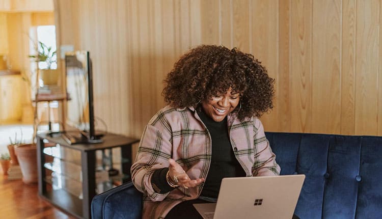 A woman sits on a blue couch, laughing and looking at her laptop on her lap.