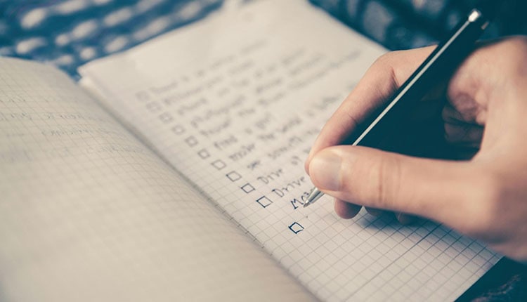 Close-up of a person's hand writing a checklist in a notebook, with checkboxes being marked off, in a monochrome tone.