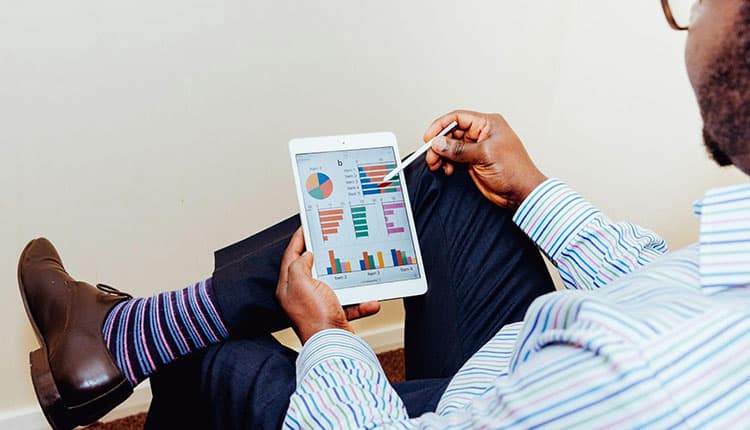 A person in business attire holds a tablet displaying charts and graphs, using a stylus to point at the screen.