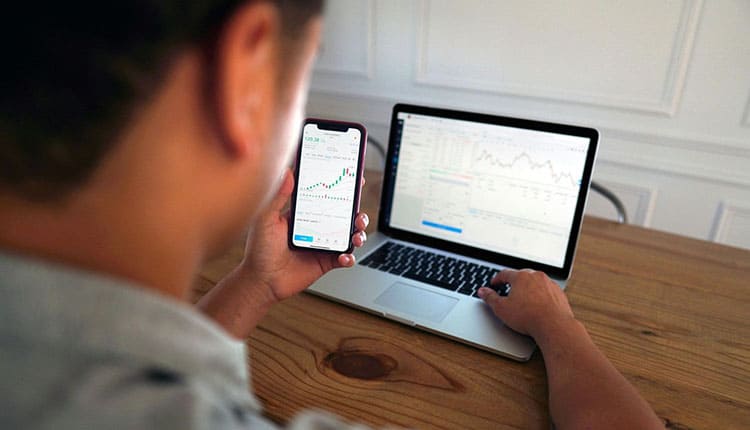 Person reviewing financial data on smartphone and laptop at a wooden desk, focusing on stock market graphs.