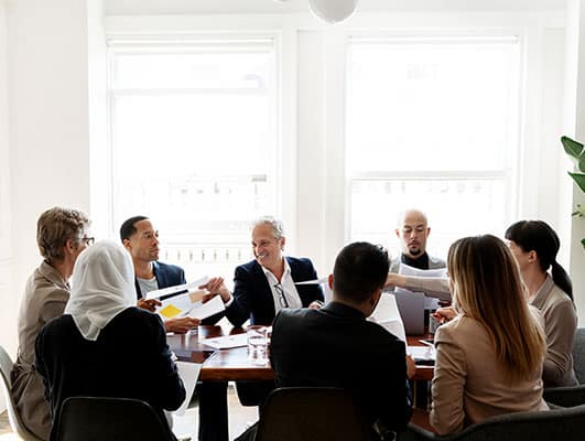 Eight people are seated around a conference table in a bright room, actively discussing and exchanging documents.