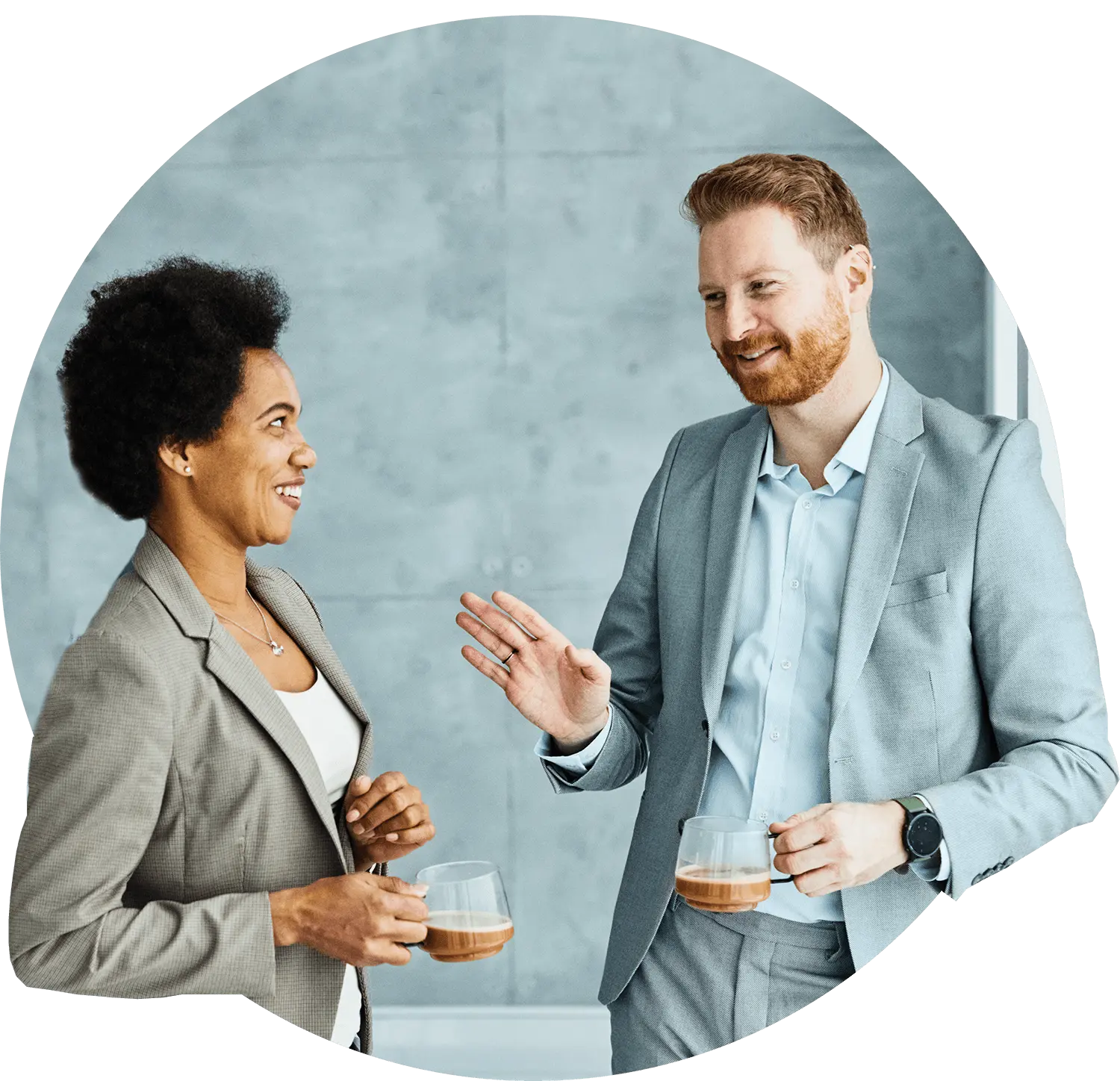 Two professionals in suits, one man and one woman, stand facing each other and smiling while holding coffee cups.