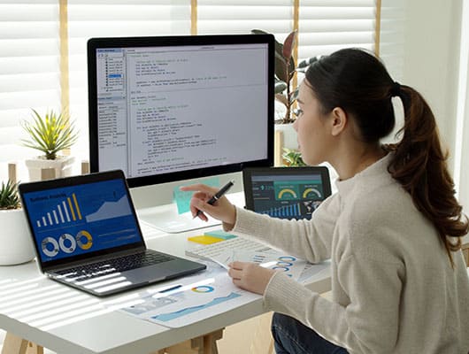 A person working at a desk with a laptop, two monitors displaying charts and code, and papers with charts.