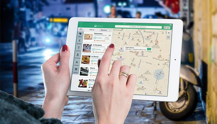 A person with red nails uses a white tablet to view a map with restaurant locations in a city street setting.