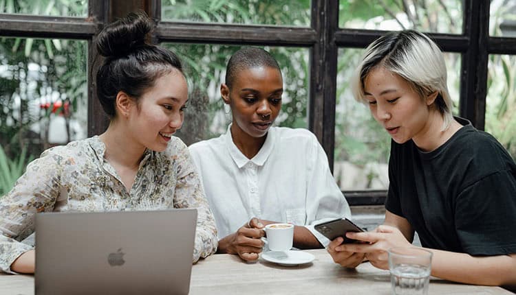 Three people are sitting together at a table. One person is using a laptop, another is holding a coffee cup, and the third is showing something on a smartphone to the others. Greenery is visible in the background.