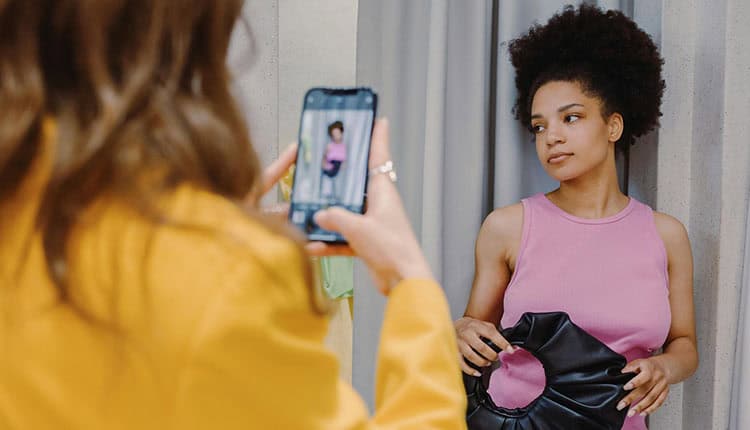 A person in a yellow outfit takes a photo of a person in a pink top holding a black bag. The person being photographed stands in front of a gray curtain.