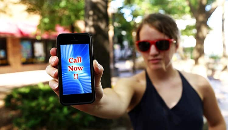 Person in sunglasses holds a smartphone displaying "Call Now!!" on the screen, standing outdoors with trees and a building in the background.