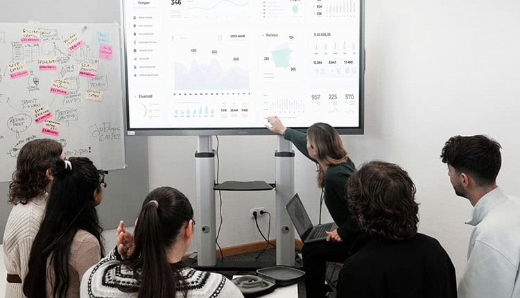 A group of people in a conference room are attending a presentation. One person is pointing at a large screen displaying data charts and graphs, while others are seated facing the screen.