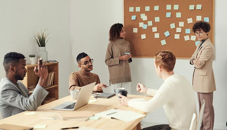 A diverse group of five people are having a discussion in a room with a bulletin board covered in sticky notes. Some are sitting at a table with laptops and notebooks, while others stand and listen.