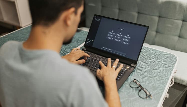 Person typing on a laptop with a dark screen interface, sitting at a table. Glasses are placed beside the laptop.