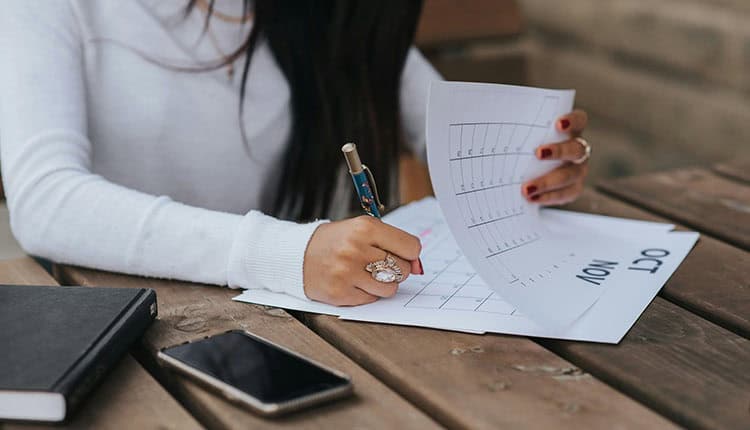 A person sitting at a wooden table, writing on a calendar. A notebook and a smartphone are placed on the table.