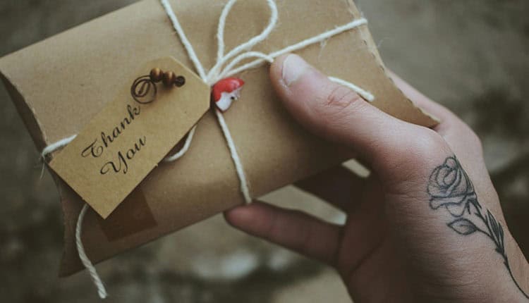 Hand holding a small brown gift box tied with string, featuring a "Thank You" tag and a rose tattoo on the wrist.