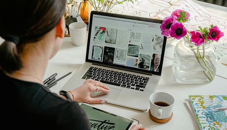 Person working at a desk with a laptop displaying design images, a tablet, coffee cup, and vase of pink flowers nearby.