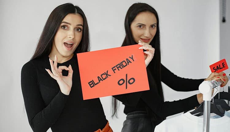 Two individuals hold red signs reading "Black Friday %" and "Sale" while standing near a clothing rack.