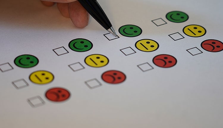 A hand holds a pen hovering over a checkbox on a survey with green, yellow, and red smiley faces arranged in rows.