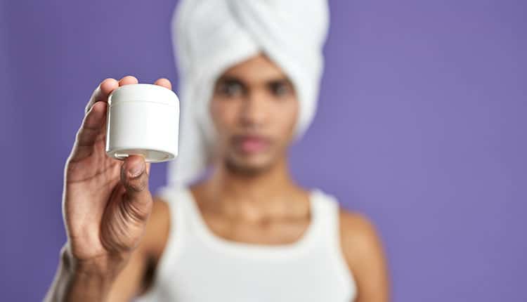 Person with a towel wrapped around their head holds up a white jar against a purple background.