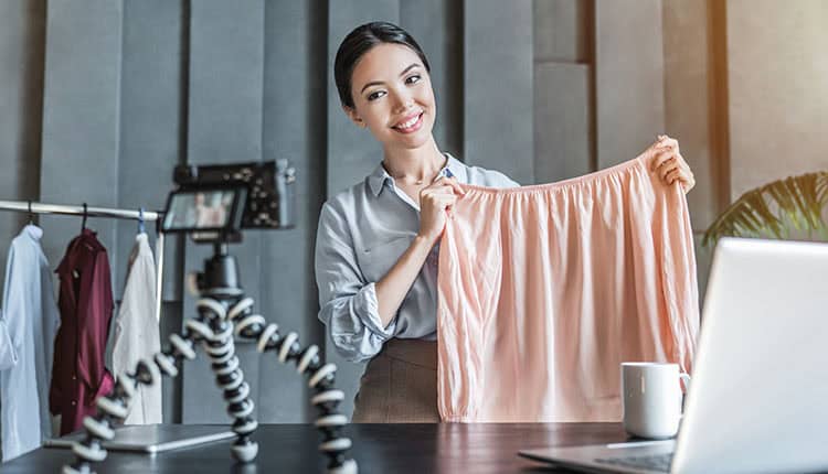 A woman holds a blouse while standing in front of a camera on a tripod. A laptop and coffee mug are on the table in front of her.