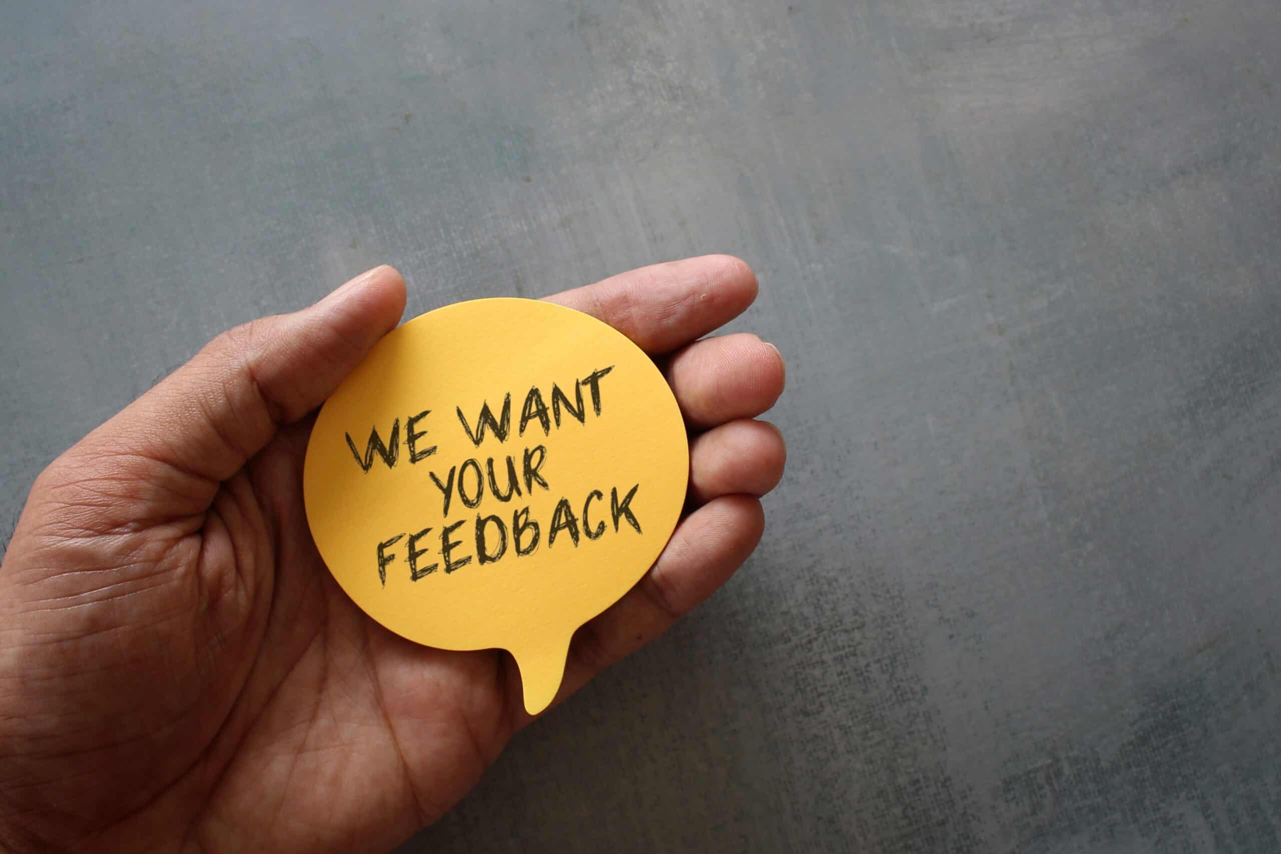 A hand holds a yellow speech bubble note with the text "We want your feedback" on a gray background.