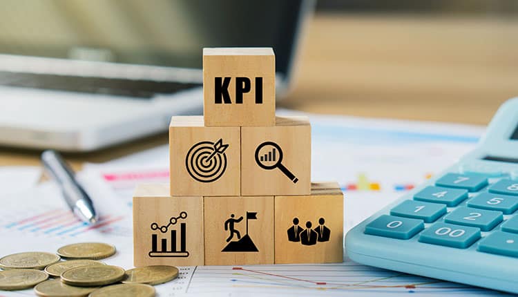 Wooden blocks labeled "KPI" with icons for target, growth, and teamwork stacked on a desk with coins, a calculator, and a laptop in the background.