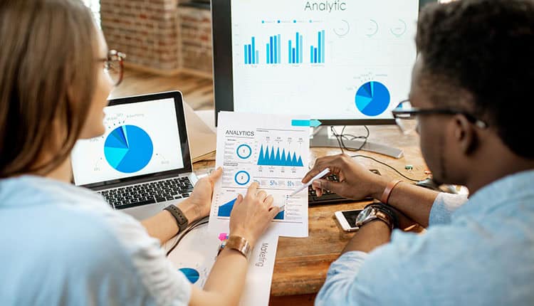 Two people analyze data charts on paper and computer screens, highlighting analytics and graphs in a workspace setting.