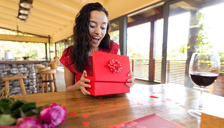 A woman opening a red gift box at a table, surrounded by rose petals and a glass of wine.