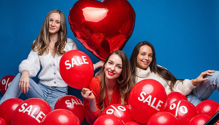 Three women pose with red balloons labeled "SALE" and a heart-shaped balloon against a blue background.