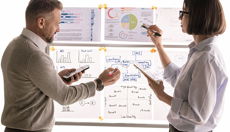 Two people discussing charts and graphs on a whiteboard. They are holding markers and a clipboard, surrounded by printed data sheets.