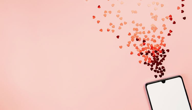 Smartphone on pink surface with heart-shaped confetti emerging from its top, creating an impression of flowing hearts.