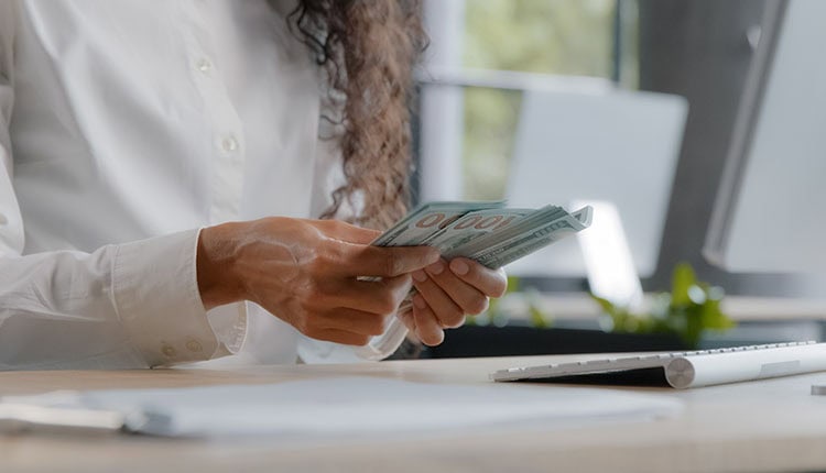 Person counting a stack of hundred-dollar bills at a desk with a computer keyboard nearby.