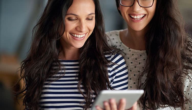 Two women with long hair are smiling while looking at a tablet. One is wearing a striped shirt, and the other a polka-dotted sweater.