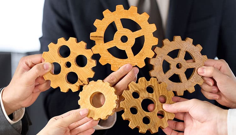 Five hands hold interlocking wooden gears, symbolizing teamwork and collaboration, against a background of a person in business attire.