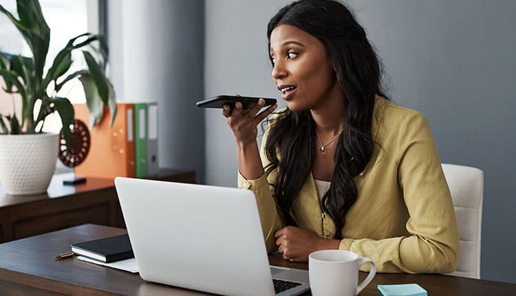 Woman in yellow blouse using smartphone for voice input at a desk with a laptop, plant, mug, and binders.