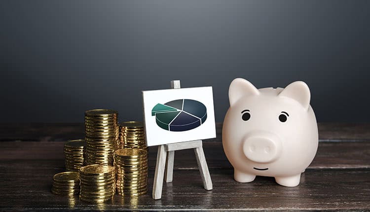 A stack of coins, a small easel with a pie chart, and a piggy bank are placed on a wooden surface.