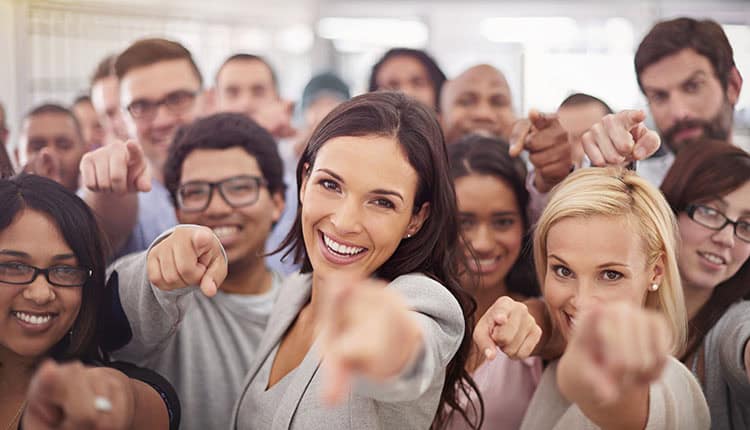 A diverse group of people standing closely together, smiling, and pointing directly at the camera.