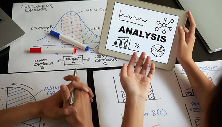 Two people discuss data charts, graphs, and handwritten notes on paper, while a tablet displays the word "Analysis" with related icons and graphs.