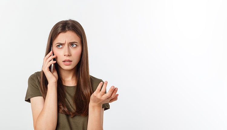 Young woman with long brown hair holding a phone to her ear, looking confused and raising her hand, standing against a plain white background.