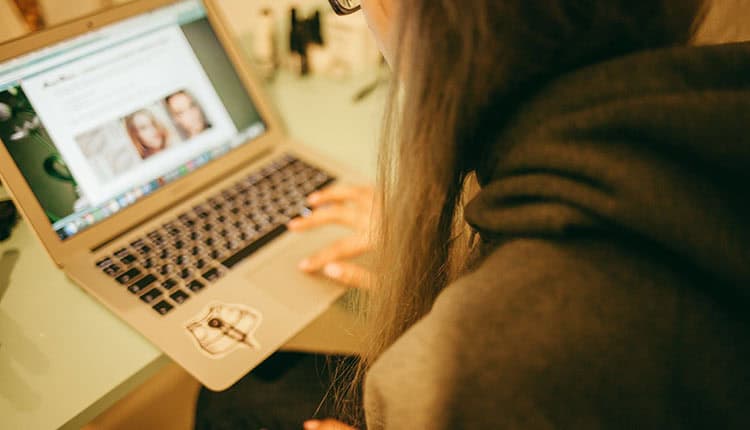 Person wearing glasses and a hoodie uses a laptop, browsing a website with images of faces displayed on the screen.