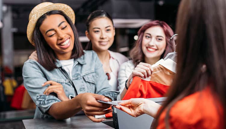 Three women stand at a store counter, one handing a credit card to a cashier while the others smile and hold clothes on hangers.