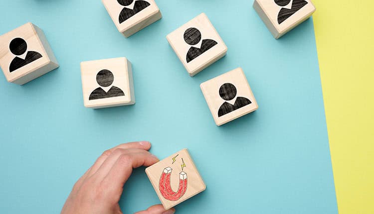 A hand holds a wooden block with a magnet illustration among blocks showing generic human icons on a blue and yellow background.