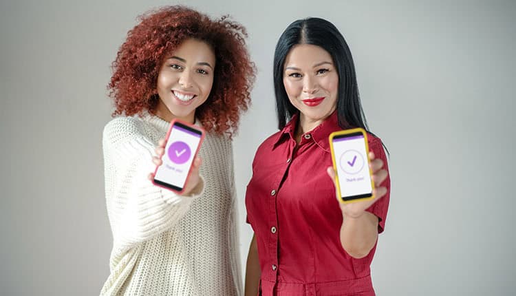 Two women standing and smiling at the camera, each holding a smartphone displaying a checkmark and the words "Thank you!" on the screen.