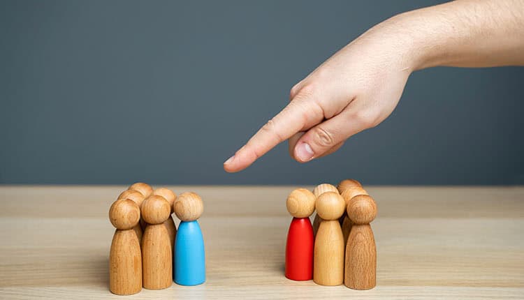 A hand points at two groups of wooden figures on a table, each group consisting of four figures, with one blue and one red figure in each group.