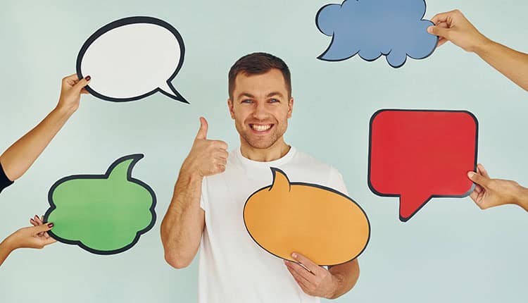 A man smiles and gives a thumbs up while surrounded by hands holding various colorful speech bubble cutouts.