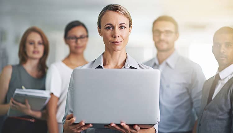 A woman holding a laptop stands in the foreground, with four colleagues blurred in the background in an office setting.