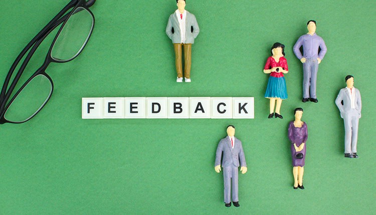 Six miniature figurines stand around the word "FEEDBACK" spelled out in letter tiles on a green background, with a pair of glasses in the upper left corner.