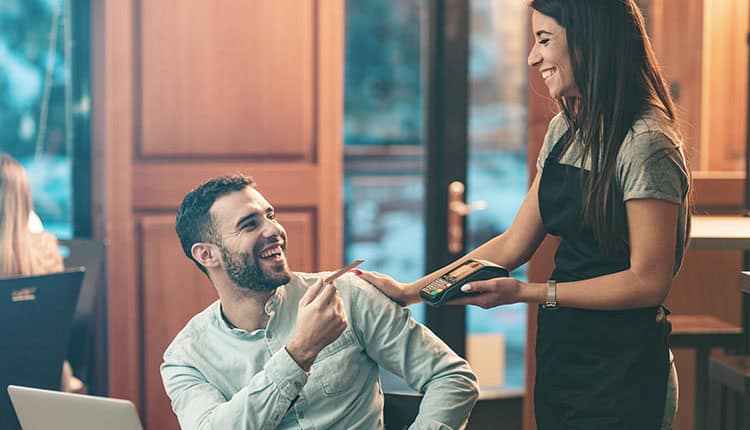 A man seated at a table hands his credit card to a smiling waitress holding a payment terminal in a restaurant.