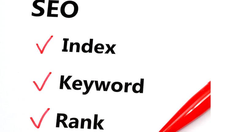 A checklist with the words "SEO," "Index," "Keyword," and "Rank," each with a red checkmark next to them and a red pen visible at the bottom.