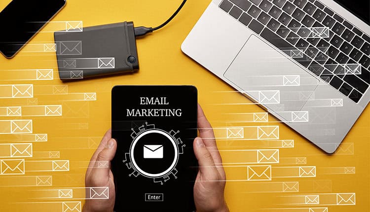 Person holding a tablet displaying "Email Marketing" near a laptop and hard drive on a yellow desk, with illustrated email icons in motion across the image.